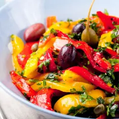 Insalata colorata di peperoni in agrodolce, con olive nere, capperi e prezzemolo fresco, servita in una ciotola blu.