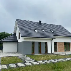 Nowy dom z szarym dachem i białymi ścianami, gotowy do odbioru. Wokół ścieżki z kostki brukowej i trawa.