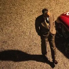 Mężczyzna w garniturze stoi obok czerwonego Ferrari. Zapowiedź filmu "Ferrari" budzi emocje, jak te wyścigi.