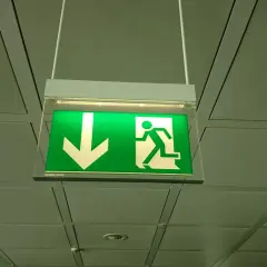 Zielone oświetlenie ewakuacyjne z ikoną strzałki w dół i biegnącą postacią, wiszące pod sufitem.