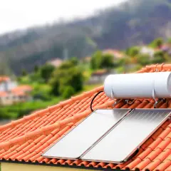 Dwa panele kolektorów solarnych i bojler na dachu z pomarańczowej dachówki, w tle zielone wzgórza i zabudowania.