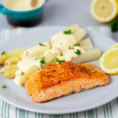 Ein saftiges Stück Lachs liegt neben weißem Spargel mit Sauce Hollandaise auf einem Teller. Garniert mit Petersilie und Zitronenscheiben.