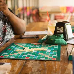Gra planszowa dla 2 osób, Scrabble, na drewnianym stole. Litery ułożone na planszy, napój i kubek w tle.