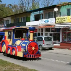 Kolorowa kolejka turystyczna jedzie ulicą. W tle sklepy, jeden z nich reklamuje "autobus 870 Gdańsk cennik".