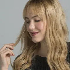 Mujer con cabello rubio ondulado y flequillo, tocando un mechón. El matizar el pelo realza su brillo y tono natural.