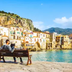 Coppia seduta su una panchina, ammira il mare e le case colorate di Cefalù, un luogo perfetto per vacanze in Sicilia sul mare.
