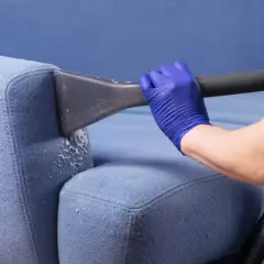 Rękawiczka w niebieskiej rękawiczce używa odkurzacza do czyszczenia niebieskiej kanapy. Dowiedz się, jak wyczyścić kanapę sodą oczyszczoną.