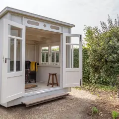 Eine charmante Gartenlaube mit offenen Türen, bereit, mit gemütlichen Möbeln eingerichtet zu werden. Ein Hocker und eine Decke deuten auf eine entspannte Atmosphäre hin.