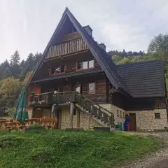 Drewniana bacówka z tarasem i schodami, otoczona zielenią i lasem.