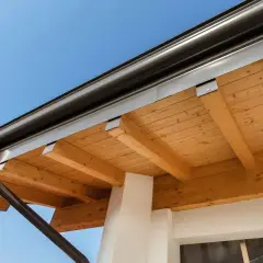 Drewniana podbitka dachu z widocznymi belkami i rynną, na tle błękitnego nieba.