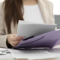 Kobieta w beżowej marynarce przegląda dokumenty w fioletowej teczce. To ważna delegacja w Niemczech, przygotowuje się do spotkania.
