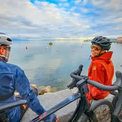 Two cyclists enjoy a break during bike guided tours in the Mediterranean, with boats on the calm sea and a coastal town in the background.