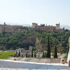Una mujer contempla la Alhambra, un lugar imprescindible que ver en Granada. El palacio se alza majestuoso sobre un monte verde.
