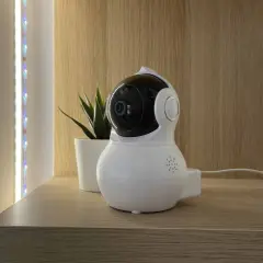 Cámara de vigilancia blanca con lente negra, parte de un sistema domótico, junto a una planta y luces LED.