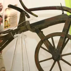 Ein Mann betrachtet das erste Fahrrad der Welt, ein hölzernes Gefährt ohne Pedale, das an ein Laufrad erinnert.