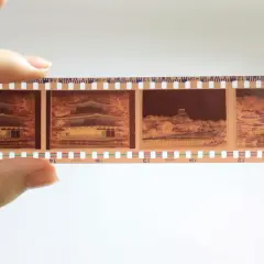 En hand håller upp en filmremsa med fyra bilder från en analog kamera. Bilderna visar en vacker byggnad med japansk arkitektur, omgiven av höstfärger.