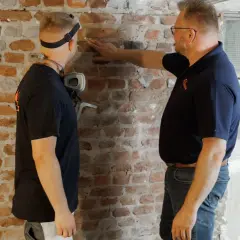 Zwei Männer untersuchen eine Ziegelwand im Keller. Einer berührt die Wand, der andere trägt eine Maske. Sie wollen die Feuchtigkeit im Keller messen.