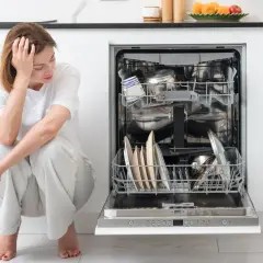 Zmartwiona kobieta siedzi obok otwartej zmywarki. Czy zmywarka ma bezpiecznik? Nie widać go, ale jest pełna naczyń.