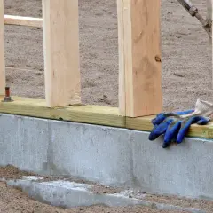 Fundament pod dom drewniany w budowie. Betonowy cokół, drewniane słupki i leżące na belce roboczej niebieskie rękawice budowlane.