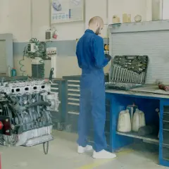 Mechanik w niebieskim kombinezonie przy silniku samochodu. Obok skrzynka z narzędziami, płyny i kanistry. Unikaj mandatu za brak przeglądu, dbaj o auto.