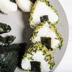 Triangular sushi rice balls, some wrapped in nori, others coated in sesame seeds and herbs, are artfully arranged on a grey plate.