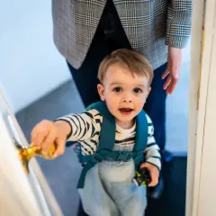 Mały chłopiec z plecakiem sięga po klamkę, a obok stoi rodzic wiodący.