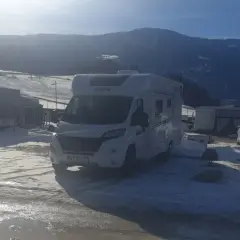 Kampery i przyczepy kempingowe na śniegu, gotowe na całoroczny camping w górach. Słońce przebija się przez chmury, oświetlając zimowy krajobraz.