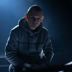 Młody polski raper w szarej bluzie z kapturem, oświetlony od tyłu niebieskim światłem.