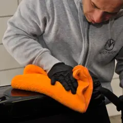 Mężczyzna w rękawiczkach czyści czarną powierzchnię pomarańczową mikrofibrą. Używa adbl apc pro do pielęgnacji wnętrza auta.
