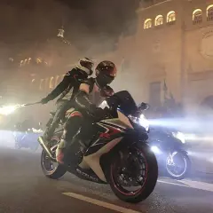Dos motoristas en una Suzuki, uno con una bengala encendida, circulan por la noche. Parece que una motocicleta puede circular sin tubo de escape, creando un espectáculo de luces y humo.