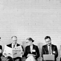 Pięciu ludzi w garniturach siedzi przed białą ceglaną ścianą. Jeden czyta gazetę, inny pracuje na laptopie. Wszyscy wyglądają jakby czekali na ważne spotkanie, może to zespół typu **ENTJ** przygotowujący się do strategicznej sesji.