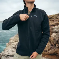 A man zips up a dark grey sweater fleece jacket, perfect for a coastal adventure. The textured fabric offers warmth and comfort.