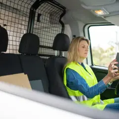 Kobieta w kamizelce odblaskowej sprawdza w telefonie DHL numer przesyłki, trzymając paczkę.