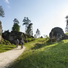 Zwei Radfahrer genießen eine Tour durch eine felsige Landschaft. Sie fahren auf einem Schotterweg, flankiert von grünen Wiesen und imposanten Felsen. Ein perfekter Tag, um in Heubach zu wandern.