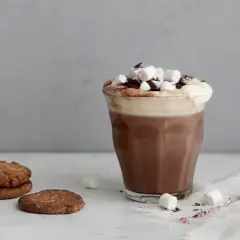 En härlig varm choklad med fluffigt skum, marshmallows och chokladströssel. Bredvid ligger några krispiga kakor.