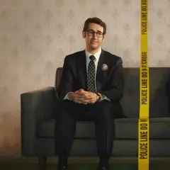 Dwóch mężczyzn siedzi na kanapie. Jeden, ubrany w garnitur, to **the good cop**. Obok niego, za taśmą policyjną, drugi mężczyzna uśmiecha się.