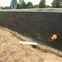 Pomarańczowa rura kanalizacyjna przechodzi przez fundament z czarnej cegły, przygotowywany do dalszych prac budowlanych.