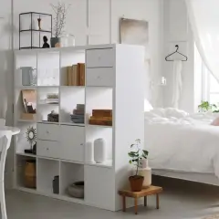 Ein heller Raum mit einem weißen Regal, das als Trenner dient. Links ein Essbereich, rechts ein Bett – ein cleverer wohn und schlafzimmer in einem raum.