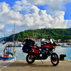 Moto rossa pronta a partire dal porto azzurro dell'Elba, con barche a vela e colline verdi sullo sfondo.