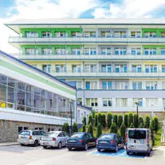 Sanatorium z rehabilitacją kręgosłupa dla rolników. Nowoczesny budynek z zielonymi elementami fasady, otoczony drzewami i parkingiem.