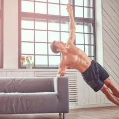 Mężczyzna ćwiczy crossfit w domu, wykonując deskę boczną na kanapie. Pokazuje, jak ćwiczyć crossfit w domu bez sprzętu.
