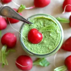 Słoik z zielonym pesto z liści rzodkiewki, ozdobiony jednym rzodkiewkowym serduszkiem. Wokół rozłożone są świeże rzodkiewki.