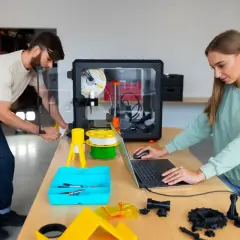 Mężczyzna i kobieta pracują przy drukarce 3D, która tworzy przedmioty. To pokazuje, czym jest drukarka 3D.