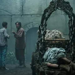 Zwei Männer diskutieren in einer mysteriösen, moosbewachsenen Kammer. Im Vordergrund schläft eine Person in einem rustikalen Bett. Szene aus "Der Greif Staffel 2".