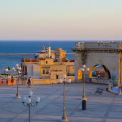 Solnedgång över Cagliari, Sardinien. En stor stenbåge badar i gyllene ljus, med havet och staden i bakgrunden.