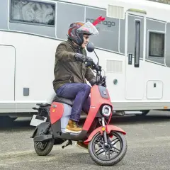 Un homme casqué conduit un scooter rouge devant un camping-car. Ce scooter est parfait pour explorer les environs lors de vos voyages en camping-car.