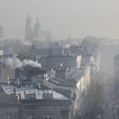 Widok na miasto w smogu z dymiącymi kominami i zarysami wież kościelnych w tle.