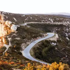Route sinueuse à travers le massif du Cap Canaille, baignée par la lumière dorée du soleil couchant. Une voiture blanche serpente sur l'asphalte.