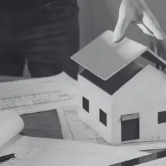 Model domu na papierach z planami. Ręka kładzie dach, symbolizując sprzeciw sąsiada na budowę lub trudności w uzyskaniu pozwolenia.