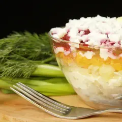 Warstwowa sałatka szuba w szklanej misie, obok pęczek koperku i widelec.